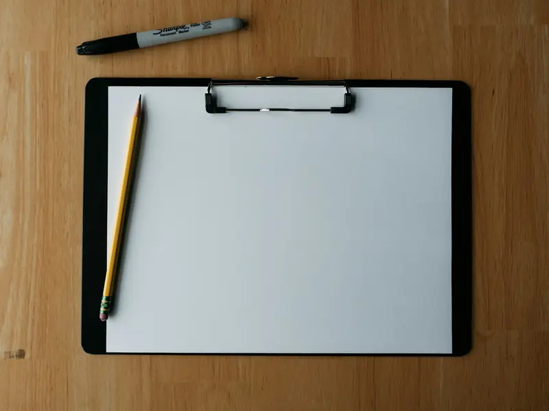 Black clipboard with blank paper, a pencil and a marker on a wood desk